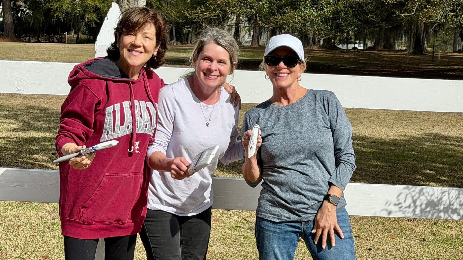 Three women volunteers smiling while painting the white fence at Coosaw Point